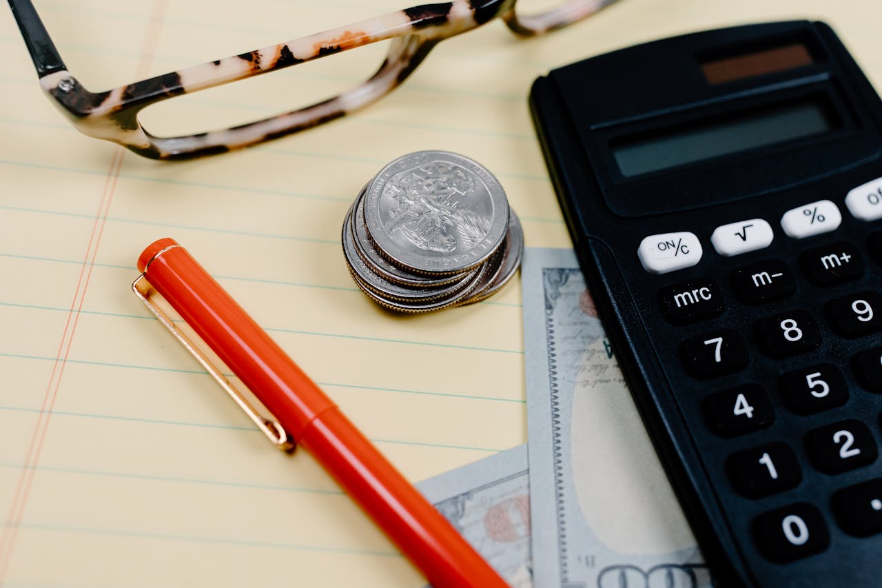 Close-up of a workspace with a calculator, coins, and glasses on a notepad.